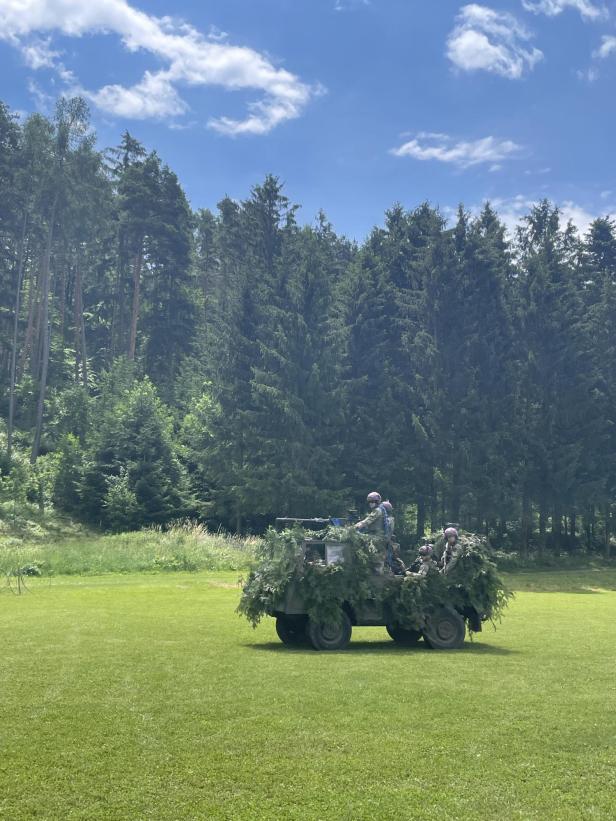 Getarnter Militärwagen mit Soldaten auf einer Wiese vor Wald.