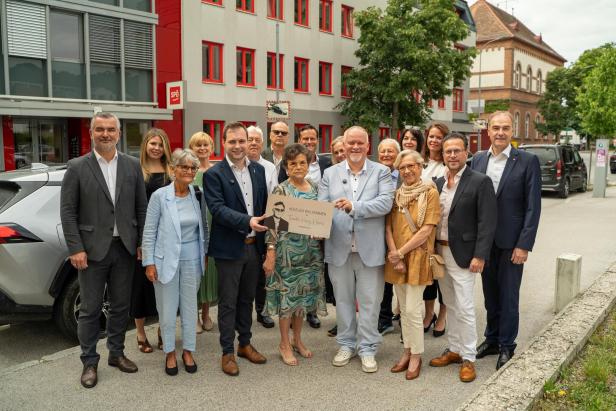Eine Gruppe von Menschen posiert vor einem Gebäude und hält ein Willkommensschild für einen Ehrengast.