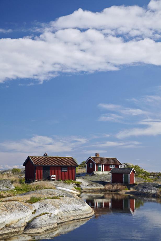 Rote Holzhütten stehen an einer felsigen Küste vor einem blauen Himmel mit weißen Wolken.