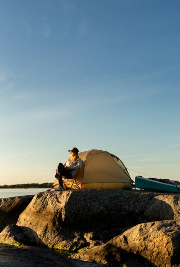 Person sitzt vor einem Zelt auf Felsen am Wasser bei Sonnenuntergang.
