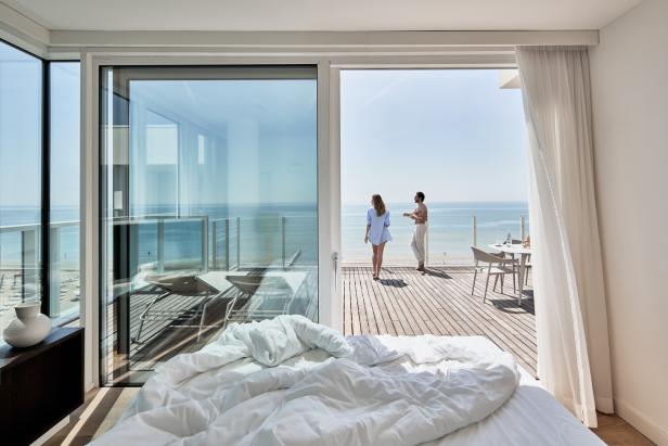 Ein Paar steht auf einem sonnigen Balkon mit Meerblick vor einem Hotelzimmer mit ungemachtem Bett.