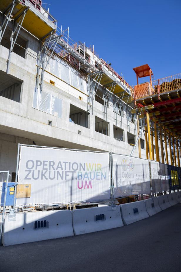 Baustelle an einem Klinikgebäude mit Gerüst und Bauzaun vor blauem Himmel.