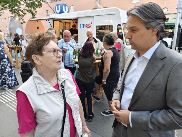 Dominik Nepp im Gespräch mit einer älteren Frau vor einem FPÖ-Infostand mit U-Bahn-Logo im Hintergrund.