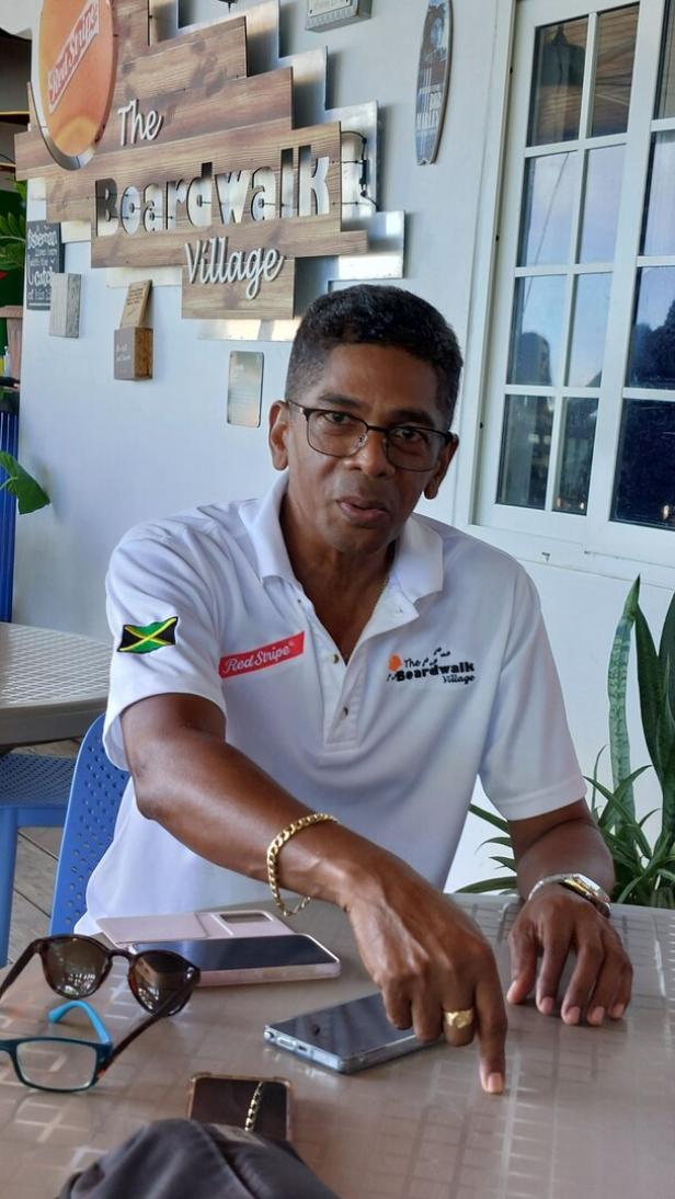 Ein Mann mit Brille und weißem Poloshirt sitzt vor einem Schild von The Boardwalk Village und zeigt auf einen Tisch.