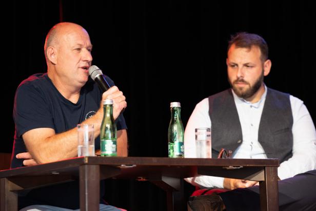 Zwei Männer sitzen an einem Tisch, einer spricht ins Mikrofon, der andere hört zu.