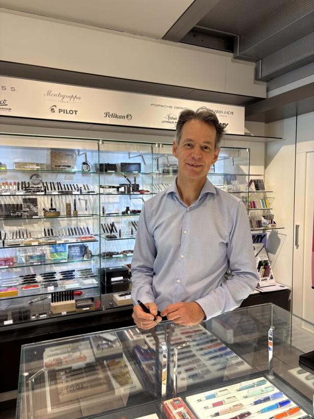 Ein Mann steht in einem Schreibwarengeschäft hinter einer Vitrine mit vielen Füllern und hält einen Stift.