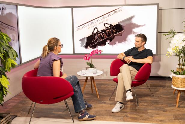 Zwei Personen sitzen sich in einem Studio auf roten Sesseln gegenüber und sprechen vor einer großen Leinwand.