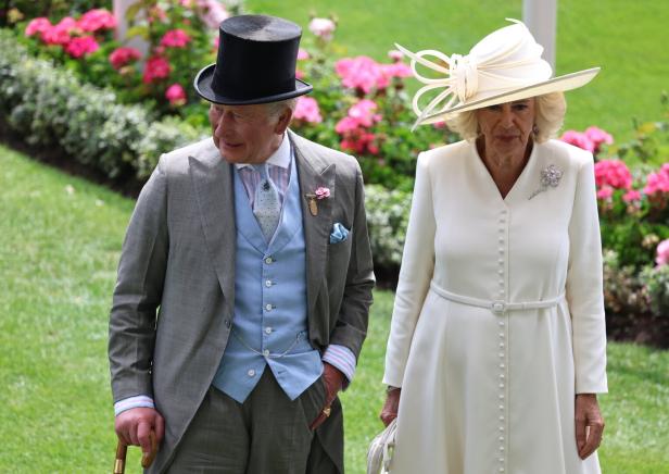König Charles III. mit Zylinder und Königin Camilla in einem weißen Kleid und Hut spazieren vor einem Blumenbeet im Garten. Charles hat die linke Hand in der Hosentasche.