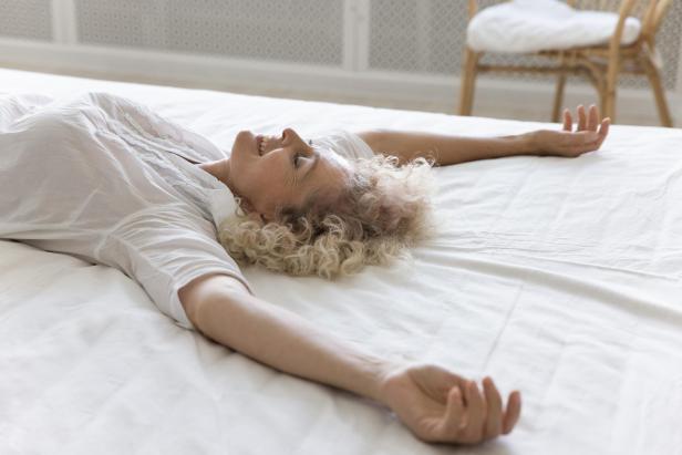 Lachende Frau mit lockigem Haar liegt entspannt auf einem weißen Bett und streckt die Arme aus.