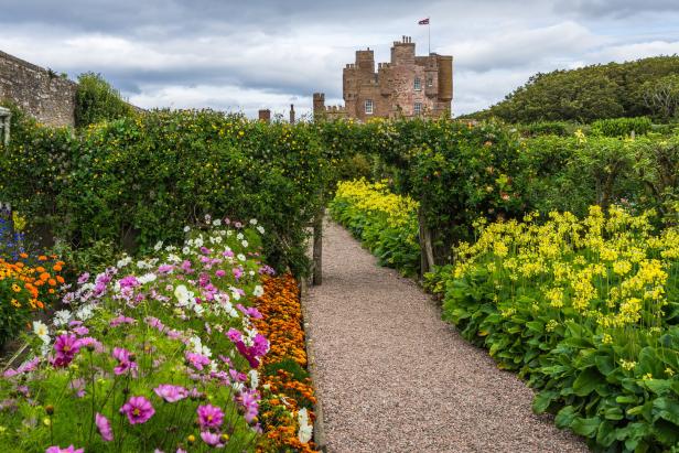 Ein Kiesweg führt durch einen blühenden Garten mit bunten Blumen auf eine historische Burg mit wehender britischer Flagge zu.