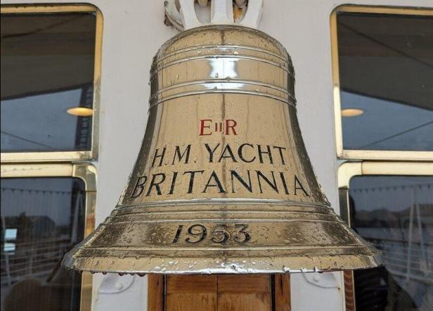 Große Schiffsglocke mit der Aufschrift „H.M. Yacht Britannia 1953“ an einer weißen Wand.