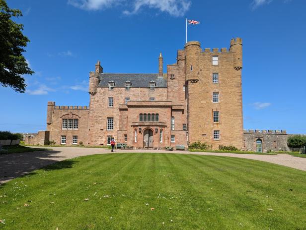 Das Castle of Mey in Schottland mit einer wehenden britischen Flagge vor einem blauen Himmel und einer gepflegten Rasenfläche.