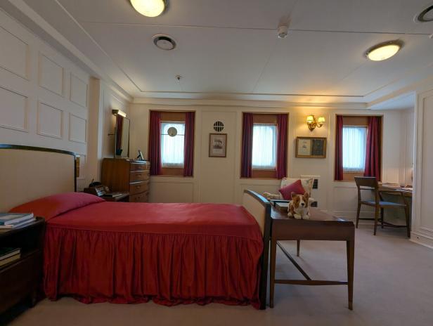 Ein klassisch eingerichtetes Schlafzimmer mit einem Bett mit roter Tagesdecke und drei Fenstern mit roten Vorhängen.