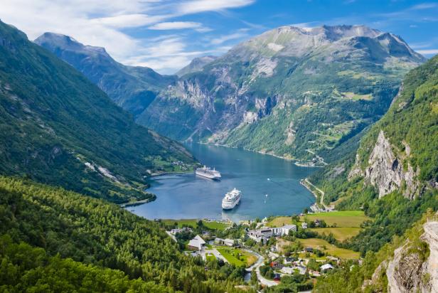 Zwei Kreuzfahrtschiffe liegen im Geirangerfjord vor der Kulisse steiler, bewaldeter Berge und dem Ort Geiranger in Norwegen.
