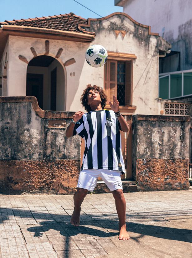 Ein barfüßiger Junge im Trikot des FC Santos jongliert vor einem alten Haus mit einem Fußball.