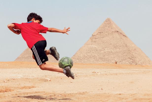 Ein Junge in rotem Trikot spielt vor den Pyramiden von Gizeh in Ägypten akrobatisch Fußball.