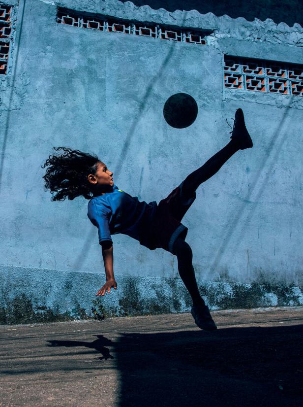 Ein Kind mit wehendem Haar führt vor einer blauen Wand einen spektakulären Fallrückzieher mit einem Fußball aus.