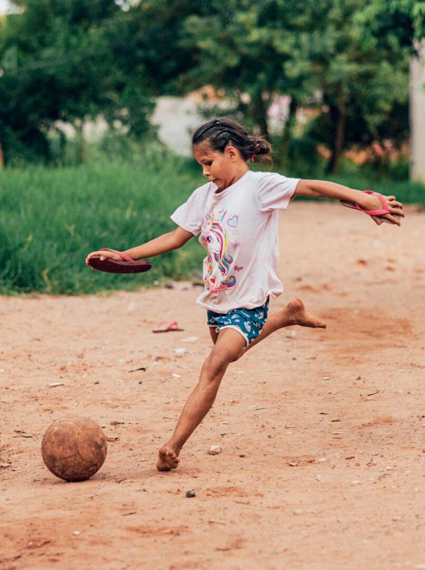 Ein junges Mädchen in einem Einhorn-T-Shirt kickt barfuß einen Fußball auf einem sandigen Weg.
