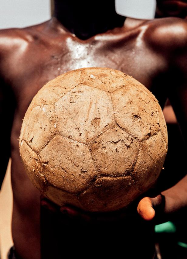 Eine Person mit verschwitztem Oberkörper hält einen stark abgenutzten, schmutzigen Fußball in den Händen.