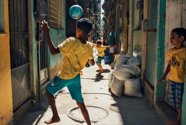 Jungen spielen in einer engen Gasse mit einem Ball, während ein Kind den Ball mit dem Kopf jongliert.
