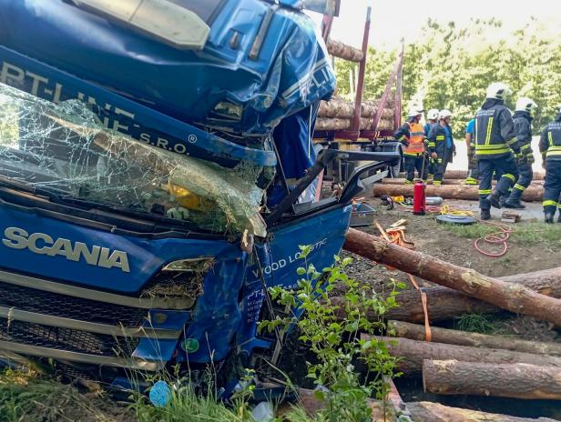 Beschädigter blauer Scania-Lkw nach Unfall mit Holzstämmen, Rettungskräfte stehen daneben.