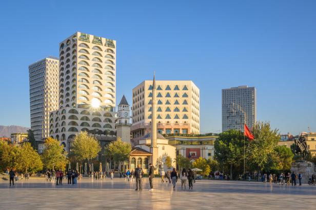 Der Skanderbeg-Platz in Tirana mit der Et'hem-Bey-Moschee, dem Uhrturm und modernen Hochhäusern unter klarem blauem Himmel.