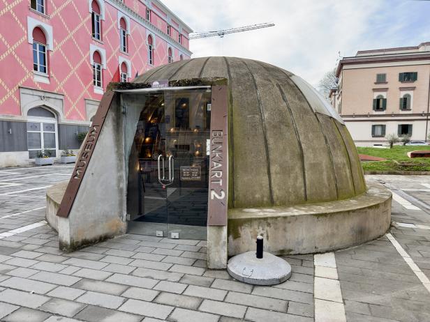 Halbkugelförmiger Bunker mit Glastür auf einem Platz, umgeben von farbigen Gebäuden.