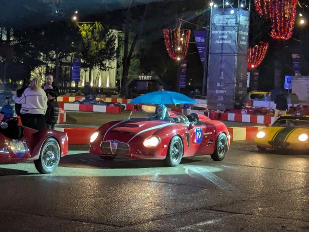 Ein roter Oldtimer mit blauem Regenschirm auf dem Dach fährt bei der Automotive Fair Albania an einer Startlinie vorbei.