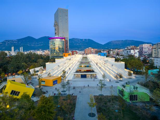 Die beleuchtete Pyramide von Tirana mit ihren begehbaren Treppen vor einer modernen Skyline und Bergen in der Abenddämmerung.