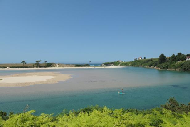 Weite Meeresbucht mit Sandbank, grünem Ufer und einer Person auf dem Stand-Up-Paddle-Board.