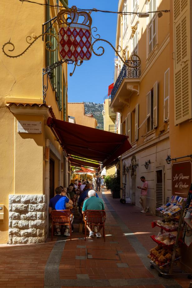 Enge mediterrane Gasse mit Straßencafé, roten Markisen und zahlreichen Sitzenden zwischen hohen Häusern.