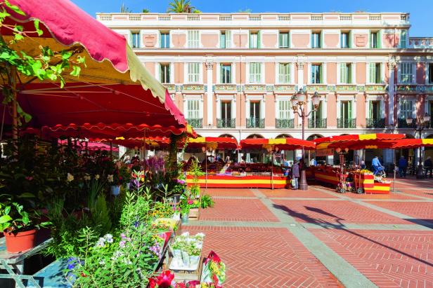 Ein sonniger Marktplatz mit rot-gelben Ständen voller Blumen und Obst vor einem historischen Gebäude mit pastellfarbener Fassade.