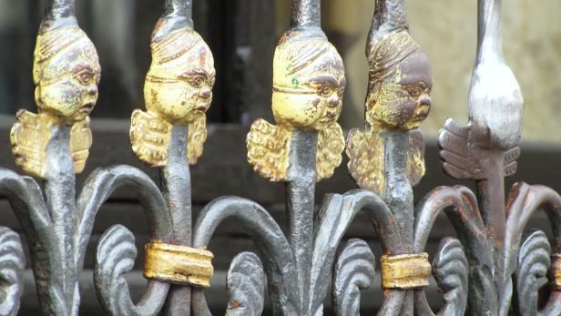 Detailansicht eines verzierten, eisernen Zauns mit Engelsgesichtern.