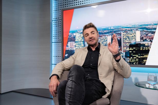 Ein Mann im hellen Blazer sitzt im Studio und spricht vor einer Stadtkulisse, die Hand erhoben.