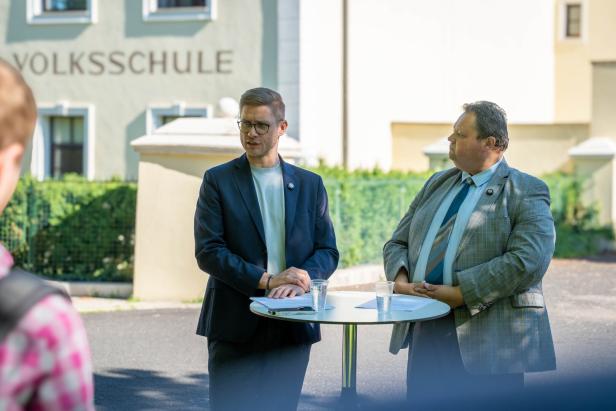 Zwei Männer im Anzug sprechen an einem Stehtisch vor einer Volksschule.