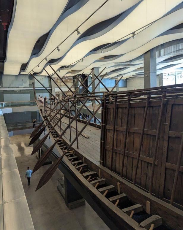 Ein historisches Holzschiff mit zahlreichen Rudern ist in einer modernen Museumshalle mit wellenförmiger Deckenkonstruktion ausgestellt.
