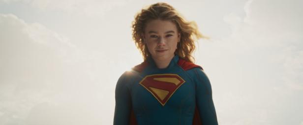 Milly Alcock als Supergirl mit blondem Haar und blauem Kostüm vor einem hellen Himmel.