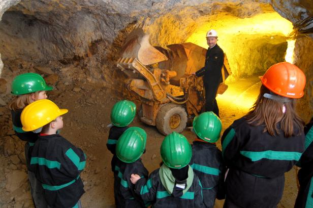 Ein Bergmann in Uniform erklärt einer Gruppe von Kindern mit Schutzhelmen in einem beleuchteten Stollen eine Bergbaumaschine.