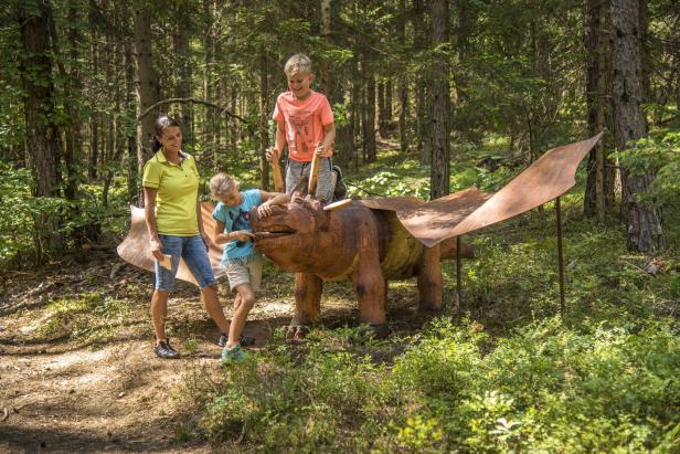 Eine Frau und zwei Kinder spielen im Wald mit einer hölzernen Drachenskulptur, die Metallflügel hat.