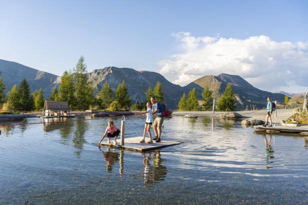 Eine Familie zieht sich auf einem Floß über einen Bergsee, während eine weitere Person am Ufer steht.