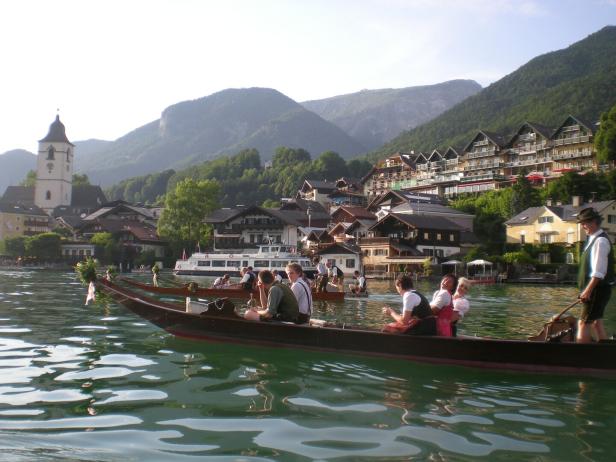 Boote mit Menschen auf einem See vor einem Bergdorf und Kirche.