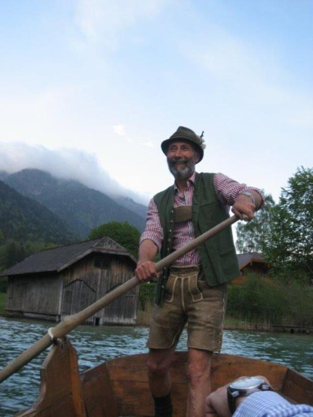 Ein Mann in Tracht rudert in einem Holzboot auf einem See vor Bergen.