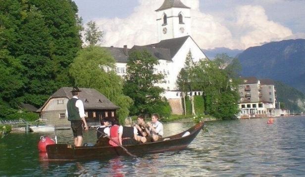 Blick über einen See auf Boote, Ufergebäude und eine weiße Kirche vor Bergen.