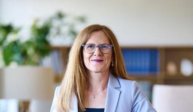 Eine Frau im hellen Blazer lehnt in einem Büro lächelnd an einem Tisch.