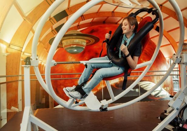 Ein junges Mädchen sitzt in einem Gyroskop-Simulator vor einer Wand mit Planetenmotiven.