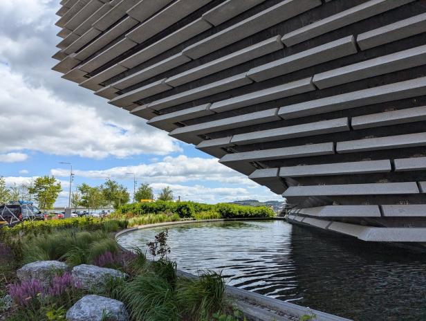 Die markante, geschichtete Fassade des V&A Dundee ragt über einem kleinen Wasserbecken mit umgebender Bepflanzung auf.