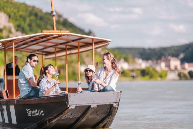Vier Personen sitzen auf einem Boot und stoßen mit Weingläsern vor einer Flusslandschaft an.