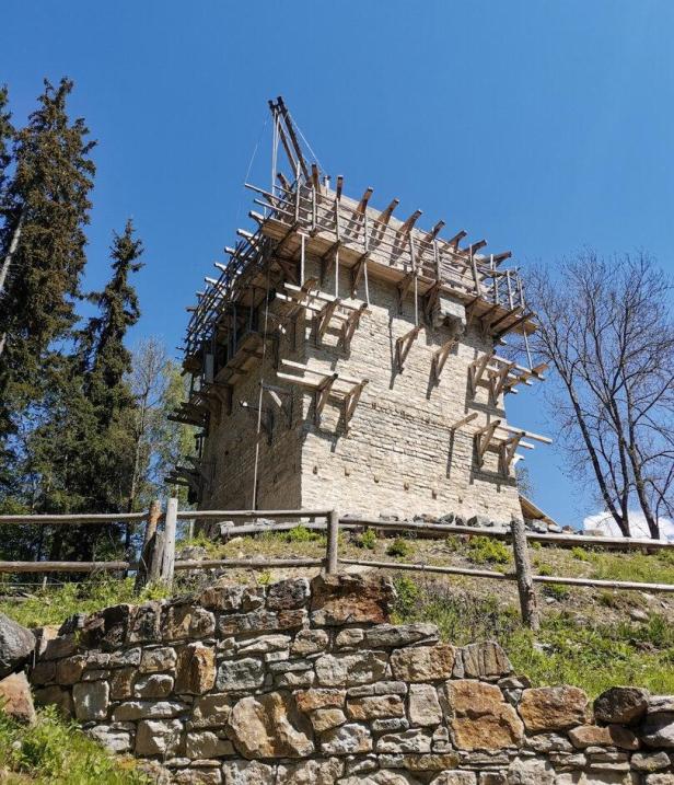 Ein Steinturm mit hölzernem Baugerüst steht auf einem Hügel vor einem klaren blauen Himmel.