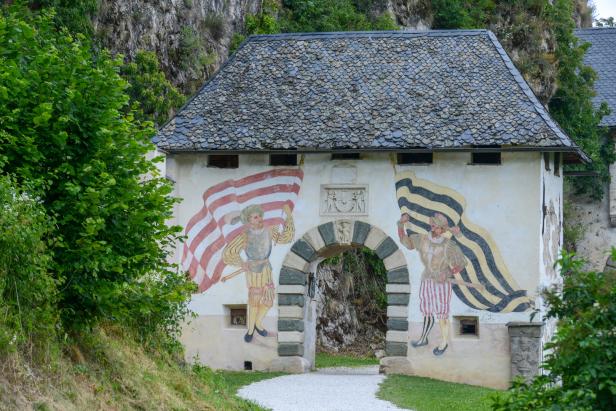 Ein historisches Torhaus mit Schiefer-Walmdach und Wandmalereien von Fahnenträgern in Renaissance-Kleidung vor einer Felswand.