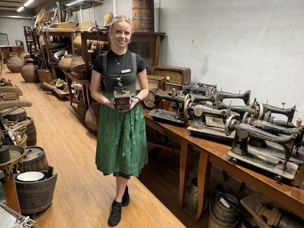 Junge Frau im Museum zwischen alten Nähmaschinen und Körben, hält ein antikes Gerät.